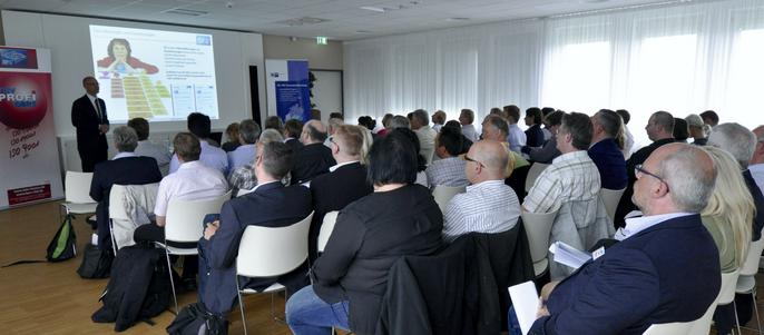 Ein Raum voller Menschen, die auf Stühlen sitzen und aufmerksam einem Vortrag zuhören. Vorne steht ein Mann, der Informationen auf einem Bildschirm präsentiert.