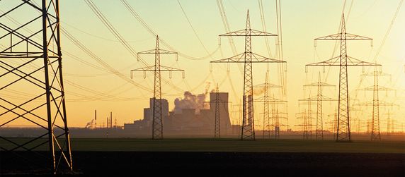 Eine weite Landschaft mit mehreren Strommasten, die durch Stromleitungen miteinander verbunden sind und zu einer weit entfernten Industrieanlage führen, aus der Rauch austritt.