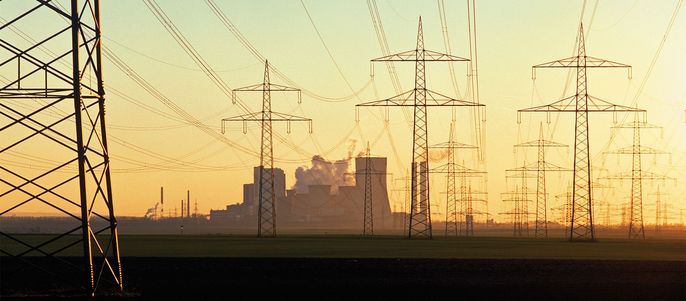 Eine weite Landschaft mit mehreren Strommasten, die durch Stromleitungen miteinander verbunden sind und zu einer weit entfernten Industrieanlage führen, aus der Rauch austritt.