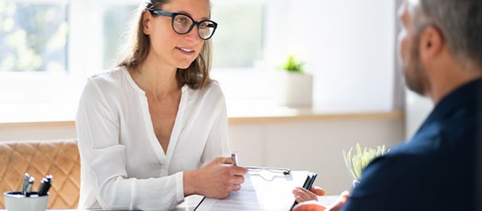 Eine Frau mit Brille und weißer Bluse, die ein Klemmbrett und einen Stift in der Hand hält, führt ein Gespräch mit einem Mann.