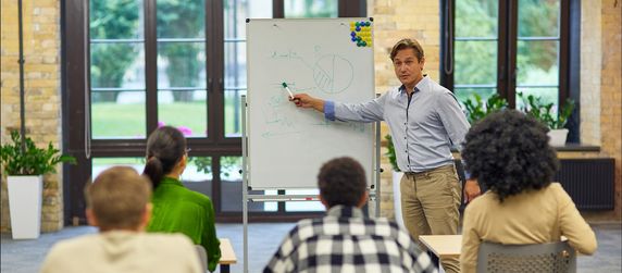 Eine Person, die etwas an ein Whiteboard zeichnet und es einigen Zuhörern erklärt.