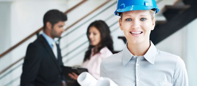 Eine Ingenieurin mit einem Schutzhelm. Im Hintergrund zwei Personen, die etwas besprechen.